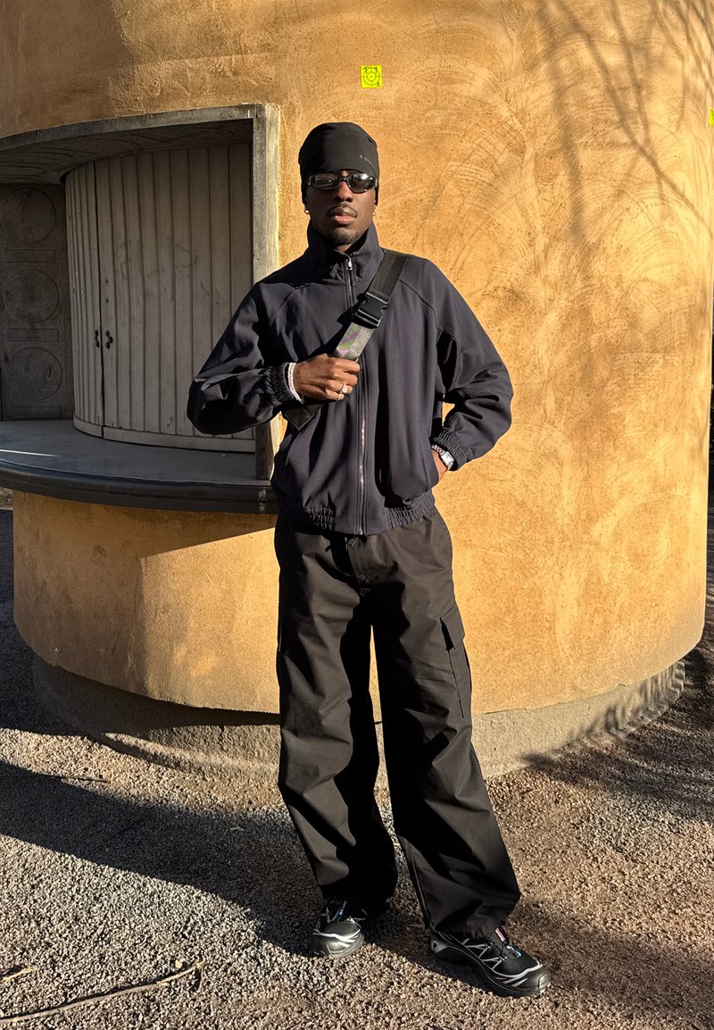 Mann med svart jakke, bukser, lue og solbriller står ute ved en teksturert buet vegg med et lukket trebordvindu.