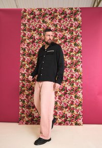 Camisa preta de botões com dois bolsos no peito, calças largas cor-de-rosa claro, e sapatos pretos. Fundo com padrão floral e acentos em rosa.