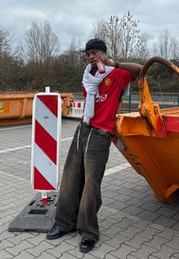 Raudona "Manchester United" marškiniai su logotipu, pilka skara, laisvi tamsūs kelnių, juodi batai. Stovi šalia stovėjimo barjero ir pramoninės konteinerio.