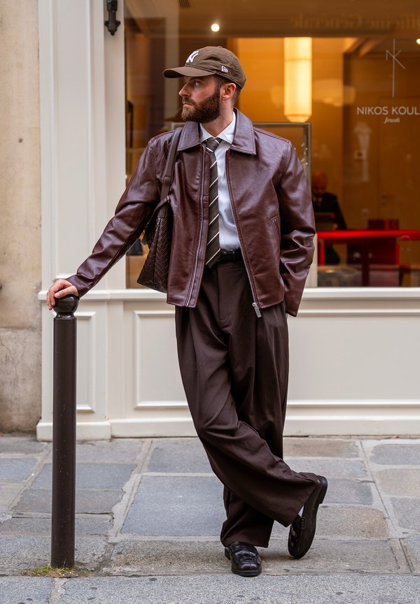 Homme en veste en cuir marron, pantalon large foncé, cravate rayée et casquette de baseball appuyé contre un poteau noir sur un trottoir urbain.