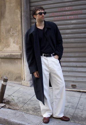 Hombre con gafas de sol negras, abrigo largo negro, camisa negra, pantalones blancos y zapatos marrones, de pie en una acera de la ciudad frente a una persiana metálica.
