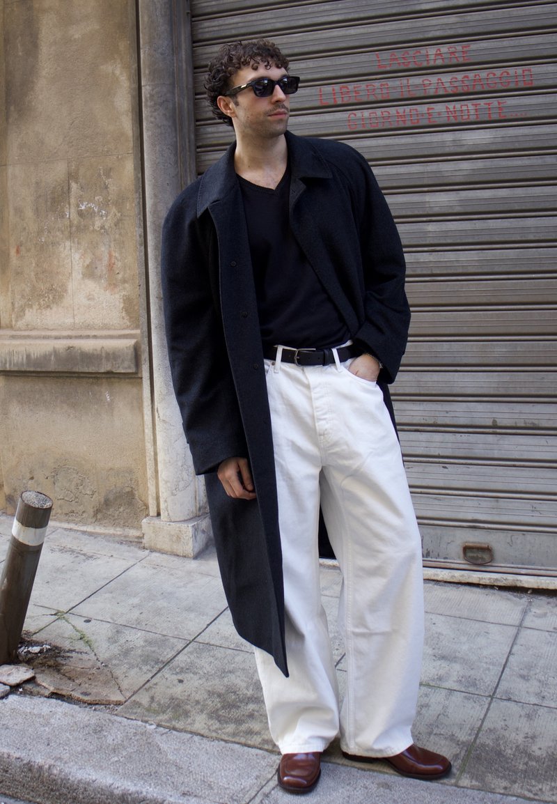 Homme portant des lunettes de soleil noires, un long manteau noir, une chemise noire, un pantalon blanc et des chaussures marron, debout sur un trottoir en ville contre un volet métallique.