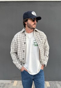 Man wearing sunglasses, navy cap, beige checked shirt over white graphic tee, and blue jeans stands with hands in pockets against grey wall.