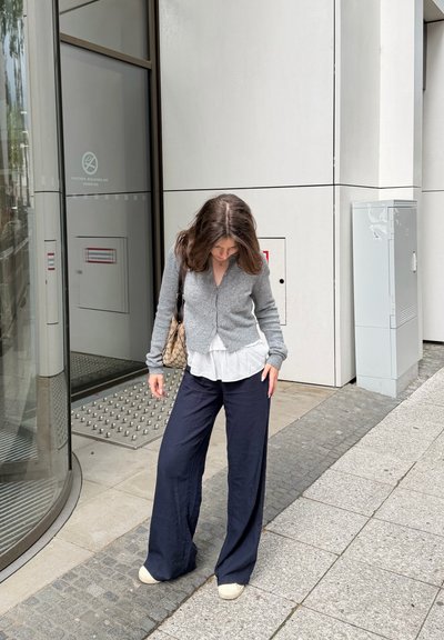 Mujer con cabello marrón que lleva un cárdigan gris, blusa blanca, pantalones anchos azul marino y zapatos beige, de pie en una acera cerca de la entrada de un edificio.