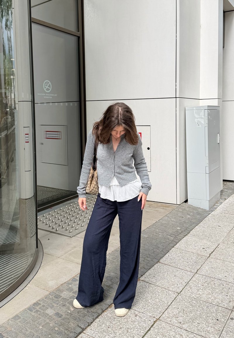 Femme aux cheveux bruns portant un cardigan gris, un chemisier blanc, un pantalon large bleu marine et des chaussures beige debout sur un trottoir près de l'entrée d'un bâtiment.