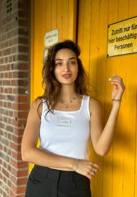 Débardeur blanc à côtes avec un patch logo, porté avec un pantalon noir taille haute. L'arrière-plan présente un mur jaune avec des panneaux et des briques.