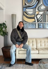 Veste bomber noire avec un col en fausse fourrure, bandoulière verte, jean bleu clair et chaussures noires. Canapé en cuir blanc en arrière-plan.