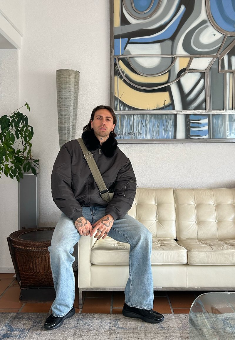 Veste bomber noire avec un col en fausse fourrure, bandoulière verte, jean bleu clair et chaussures noires. Canapé en cuir blanc en arrière-plan.