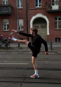 Fiatal személy fekete sportfelszerelésben és szürke sportcipőben magasra nyújtja a lábát az utcán, háttérben vörös téglás épület és kerékpárok.