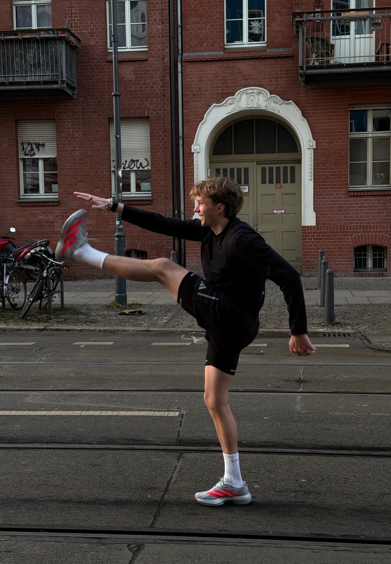 Junge Person in schwarzer Sportkleidung und grauen Turnschuhen streckt Bein hoch auf der Straße, im Hintergrund rotes Backsteingebäude und Fahrräder.