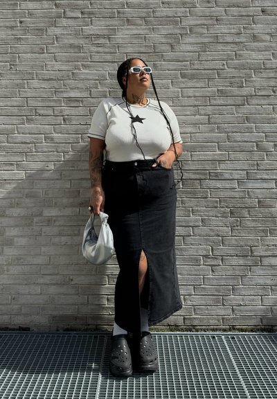 Falda maxi de mezclilla negra con una abertura frontal, camiseta blanca de canalé con una estrella negra, bolso plateado y zuecos negros con agujeros de ventilación.