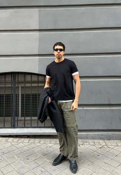 Hombre con gafas de sol negras, camiseta negra, pantalones cargo oliva, sosteniendo una chaqueta negra, de pie en una acera de baldosas junto a un edificio gris.