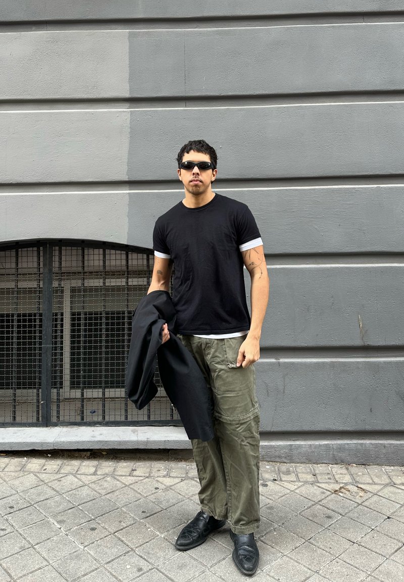 Mann mit schwarzen Sonnenbrillen, schwarzem T-Shirt, olivfarbenen Cargo-Hosen, der eine schwarze Jacke hält und auf einem gefliesten Bürgersteig neben einem grauen Gebäude steht.