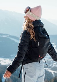 Jeune femme en tenue d'hiver avec bonnet rose et lunettes de ski, debout les bras ouverts, admirant un paysage de montagne enneigé.