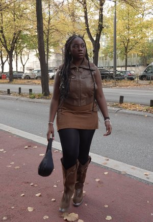 Chaleco de cuero marrón, falda corta de ante marrón claro, medias negras, botas marrones hasta la rodilla. Persona llevando un bolso negro caminando por una calle cubierta de hojas.