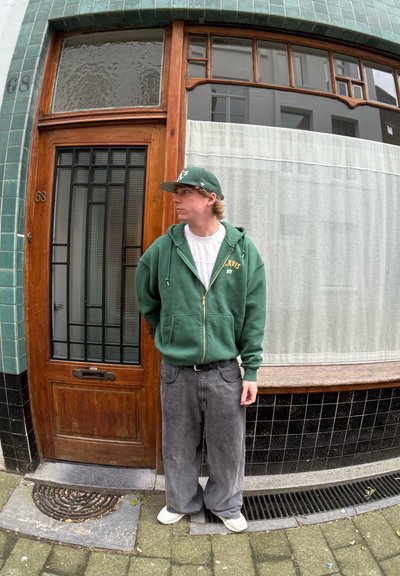 Sudadera verde con cremallera y bolsillos laterales, con letras doradas, combinada con unos vaqueros grises holgados y zapatillas blancas, de pie junto a una puerta de madera.