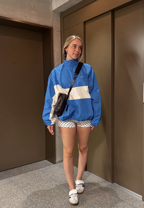 Mladá žena v modrej bunde Nike, bielych kraťasoch s bodkami, bielych teniskách a čiernej crossbody taške stojí pri hnedých dverách výťahu.