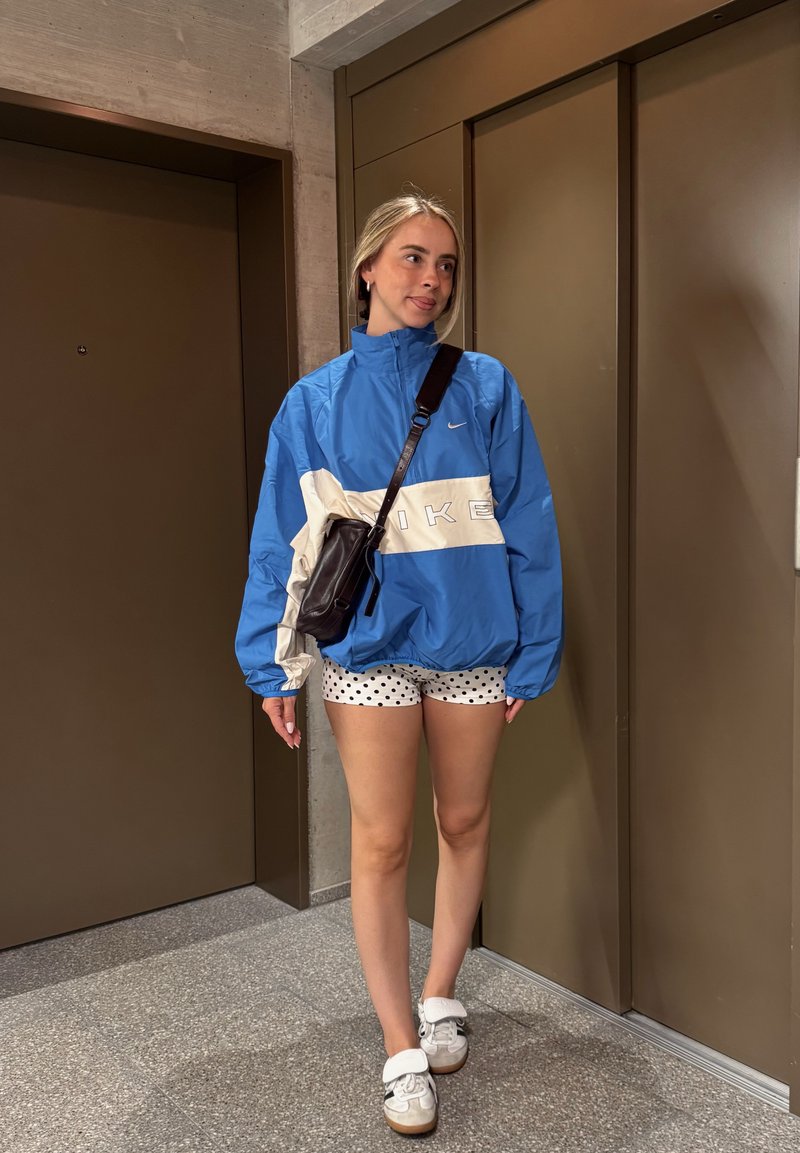 Jeune femme en veste Nike bleue, short blanc à pois, baskets blanches et sac bandoulière noir, debout près des portes marron de l'ascenseur.