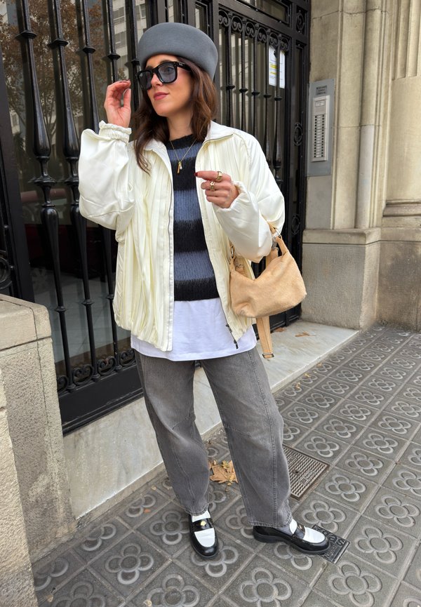 Kvinde iført grå baret, oversized hvid jakke, stribet sweater, grå jeans, sort-hvide sko og solbriller, holder en beige håndtaske på byens fortov.