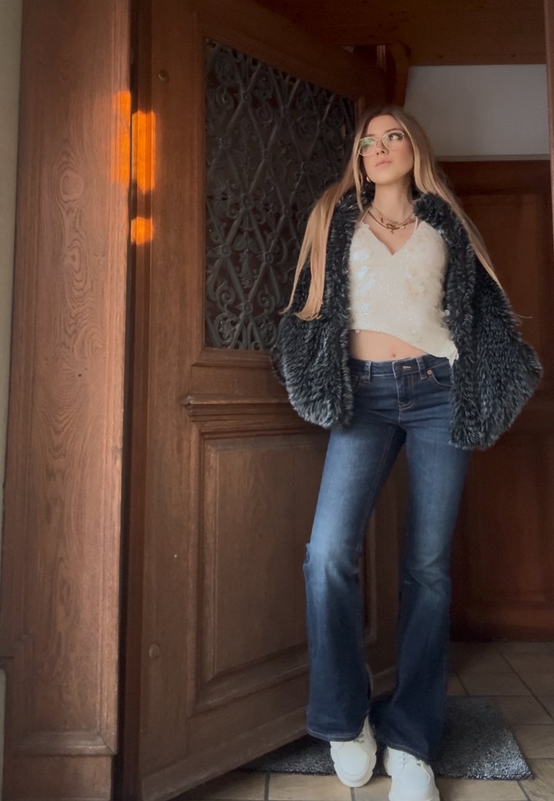 Junge Frau mit Brille, weißem Croptop, schwarzer Pelzjacke, blauen Schlagjeans und weißen Turnschuhen steht drinnen neben einer Holztür.