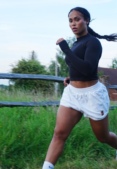 Top negro de manga larga combinado con pantalones cortos deportivos blancos. La persona está corriendo en un área de hierba con una cerca de madera al fondo.