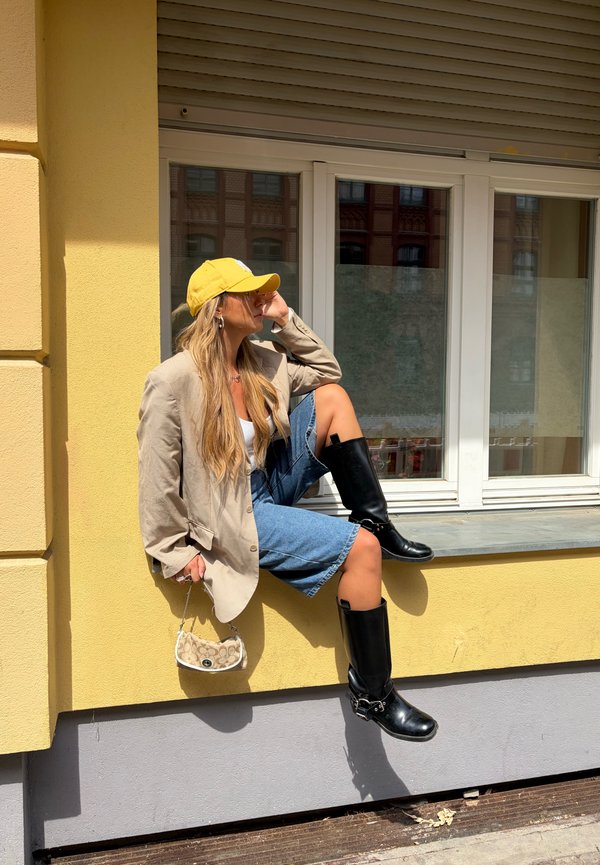 Žena s žltou čiapkou, béžovou bundou, rifľovými kraťasmi a čiernymi topánkami sedí na parapete pri žltej stene.