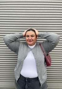 Cardigan gri, top alb și blugi negri cu talie înaltă. Mâneci scurte, închidere cu butoni la cardigan, ținând o poșetă burgundă.