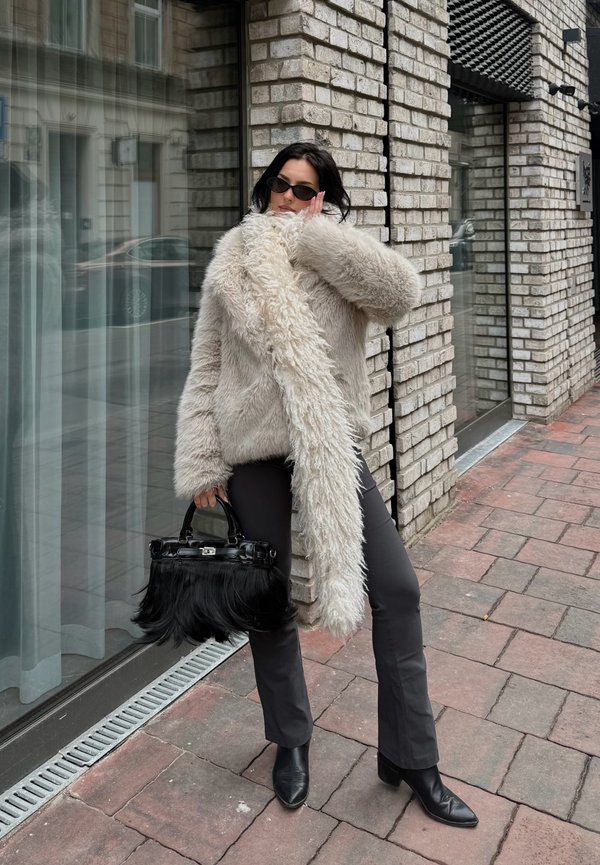 Persoon in een oversized beige bontjas, zwarte broek en laarzen staat op een bakstenen stoep en houdt een zwarte bonthaasttas vast bij een bakstenen gebouw.