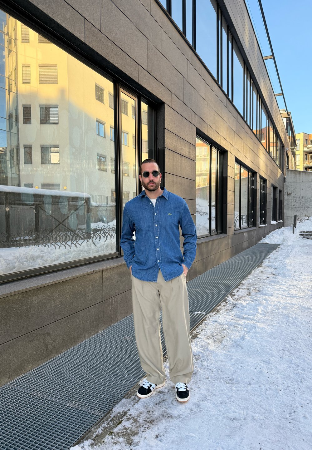Denim gombos ing, khaki nadrág, napszemüveg; áll egy havas járdán egy modern üvegezett épület mellett.