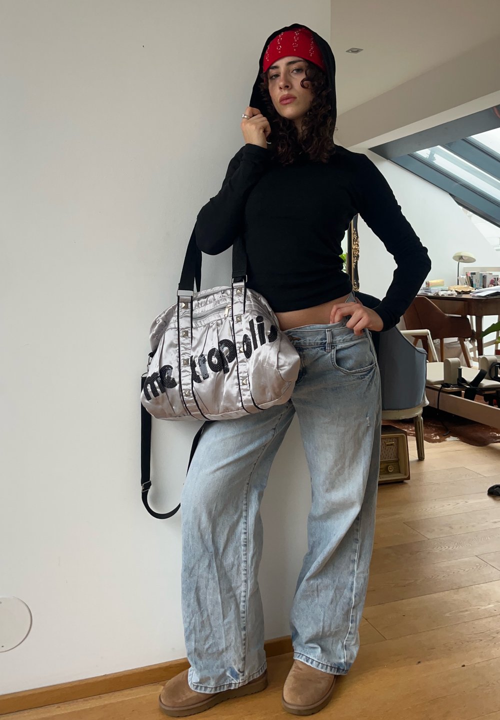Sac en satin argenté avec le texte noir "metropolis", porté à l'épaule. Haut noir à manches longues, jeans baggy en denim clair, bottes marron et bandana rouge.