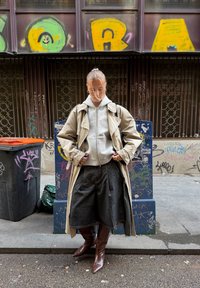 Helebeežjas beežist trents, halli kapuutsiga pusa all, kombineeritud lahtiste tumedate teksapükstega ja pruunide põlveni ulatuvate saapadega. Linnamaastik grafitiga.