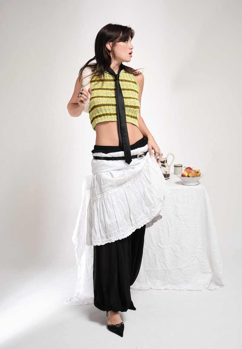 Femme en jupe noire et blanche superposée, crop top rayé jaune, cravate noire, tenant un châle, debout près d'une table avec un bol de fruits et un pichet.