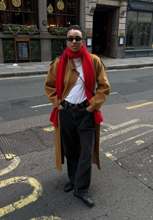 Persona de pie en la calle con abrigo marrón, bufanda roja, camisa blanca, pantalones negros, gafas de sol y zapatos negros, con las manos en los bolsillos.