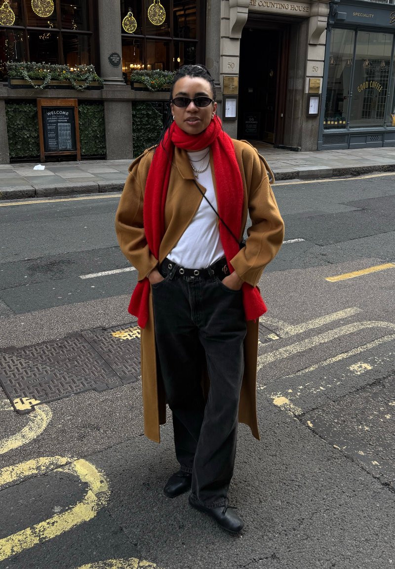 Personne debout dans la rue portant un manteau marron, une écharpe rouge, une chemise blanche, un pantalon noir, des lunettes de soleil et des chaussures noires, les mains dans les poches.