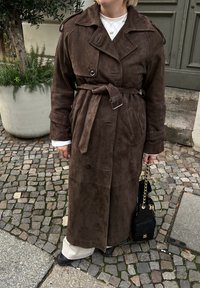 Personne portant un long trench-coat en daim marron avec ceinture nouée, pantalon blanc, chaussures noires, tenant un sac à main noir avec une lanière chaîne dorée.