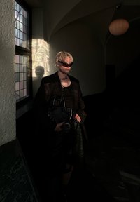 Μαύρο σακάκι κομμένο πάνω από σκούρο τοπ, συνδυασμένο με μαύρη τσάντα και γυαλιστερό κολιέ με αλυσίδα. Διαθέτει γωνιακά γυαλιά ηλίου και υφασμένο δάπεδο.