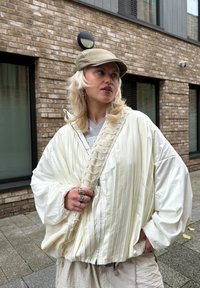 Balta, krokota jaka ar pārmēru plašām piedurknēm, kombinēta ar pelēku kreklu un bēšām biksēm. Izskatu papildina gaiši zaļa cepure un vairāki gredzeni.