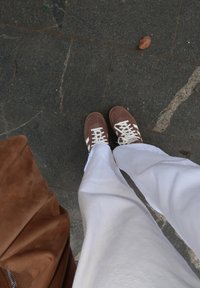 Personne portant des baskets marron et blanches et un pantalon blanc, debout sur un trottoir gris près d'un sac en daim marron et d'une feuille tombée.