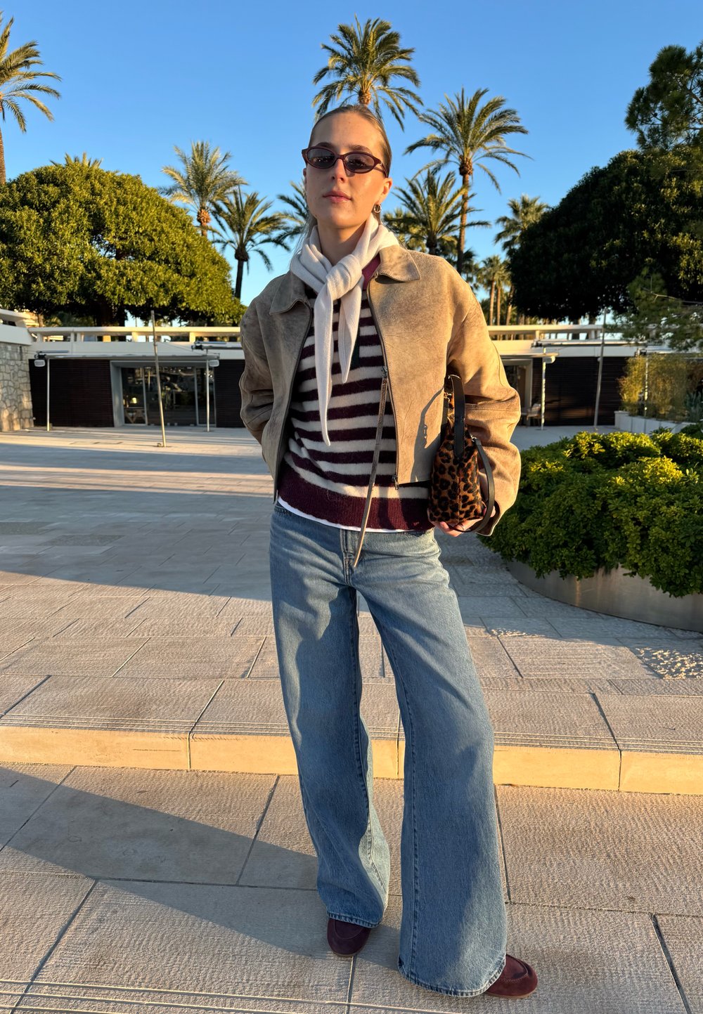 Veste en daim marron clair portée par-dessus un pull rayé bordeaux et blanc, associée à un jean bleu à jambes larges et des chaussures bordeaux. Petit sac imprimé léopard.