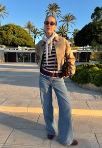 Veste en daim marron clair portée par-dessus un pull rayé bordeaux et blanc, associée à un jean bleu à jambes larges et des chaussures bordeaux. Petit sac imprimé léopard.