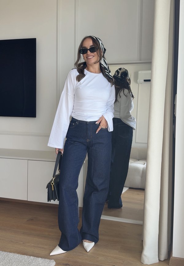 Donna con camicia bianca a maniche lunghe, jeans blu scuro, scarpe bianche appuntite, occhiali da sole neri e foulard stampato che tiene una borsa nera al chiuso.