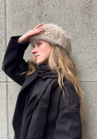 Νεαρή γυναίκα με μαύρο παλτό και κασκόλ, φορώντας ένα μπεζ γούνινο καπέλο, προστατεύει τα μάτια της ενώ στέκεται μπροστά σε ένα υφή γκρι τοίχο με νιφάδες χιονιού πάνω της.