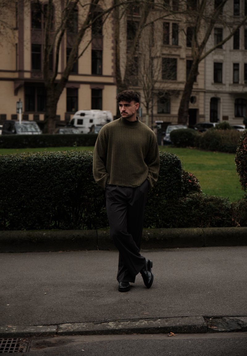 Maglione verde a coste con orlo non rifinito, abbinato a pantaloni neri e scarpe nere; in piedi davanti a uno sfondo urbano sfocato.