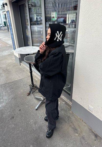 Persona con una chaqueta negra, capucha de los New York Yankees negra y pantalones a rayas parada en la acera junto a una mesa de café y una ventana.