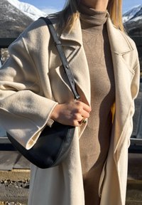 Femme portant une robe col roulé beige et un manteau crème, tenant un sac à bandoulière en cuir noir, debout en plein air avec des montagnes enneigées en arrière-plan.