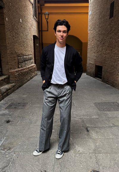 Cárdigan negro sobre una camiseta blanca, combinado con pantalones de pierna ancha grises y zapatillas negras. Callejón de adoquines con paredes de ladrillo.