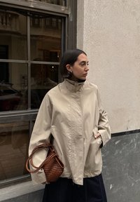 Beež suur mantel kõrge kraega ja esipocketitega, mille juurde kuulub väike punutud pruun käekott. Taustal on hall sein ja aken.