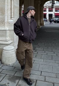 Sweat à capuche zippé marron foncé, pantalon cargo beige avec poches latérales, et bottes à plateforme marron. Une casquette grise complète ce look urbain décontracté.