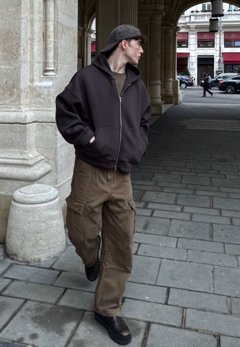 Sweat à capuche marron foncé avec fermeture éclair, pantalon cargo beige avec poches latérales, et bottes à plateforme marron. Casquette grise complète le look urbain décontracté.