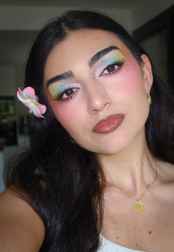Woman with long dark hair, rainbow eyeshadow, pink rhinestones around eyes, glossy brown lips, yellow flower in hair, and gold necklace and earrings.